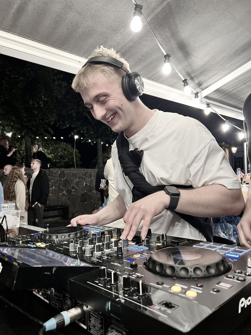 Emil Boel med høretelefoner smiler, mens han spiller ved DJ-pulten under lyskæder til en udendørs fest